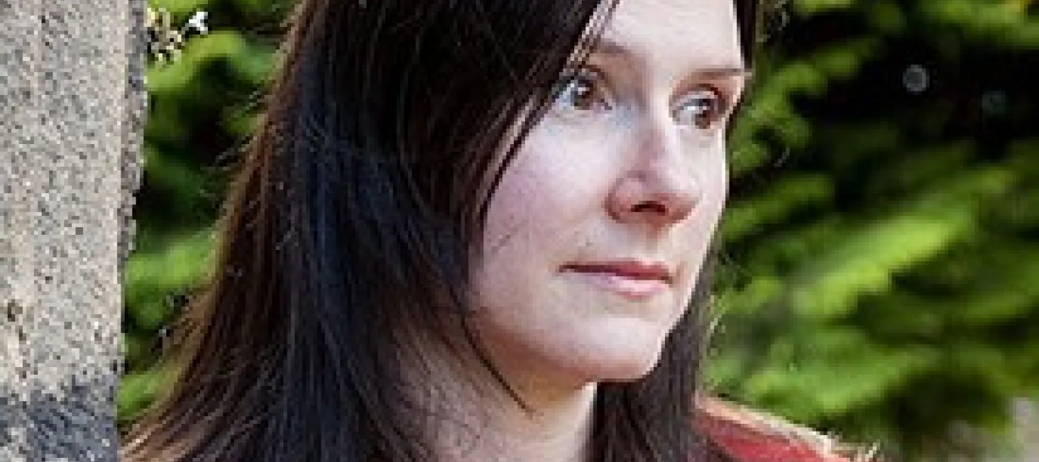Professor Fiona Edmonds has long dark hair and wears a red coat. She stands against a stone wall, gazing thoughtfully to the side. Lush green foliage forms the background.