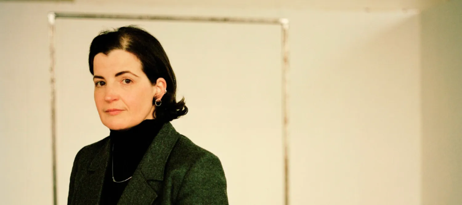 Dr Hannah Proctor has short dark hair, and wears a dark turtleneck and blazer. Dr Proctor stands indoors in front of a blank wall and looks directly at the camera.