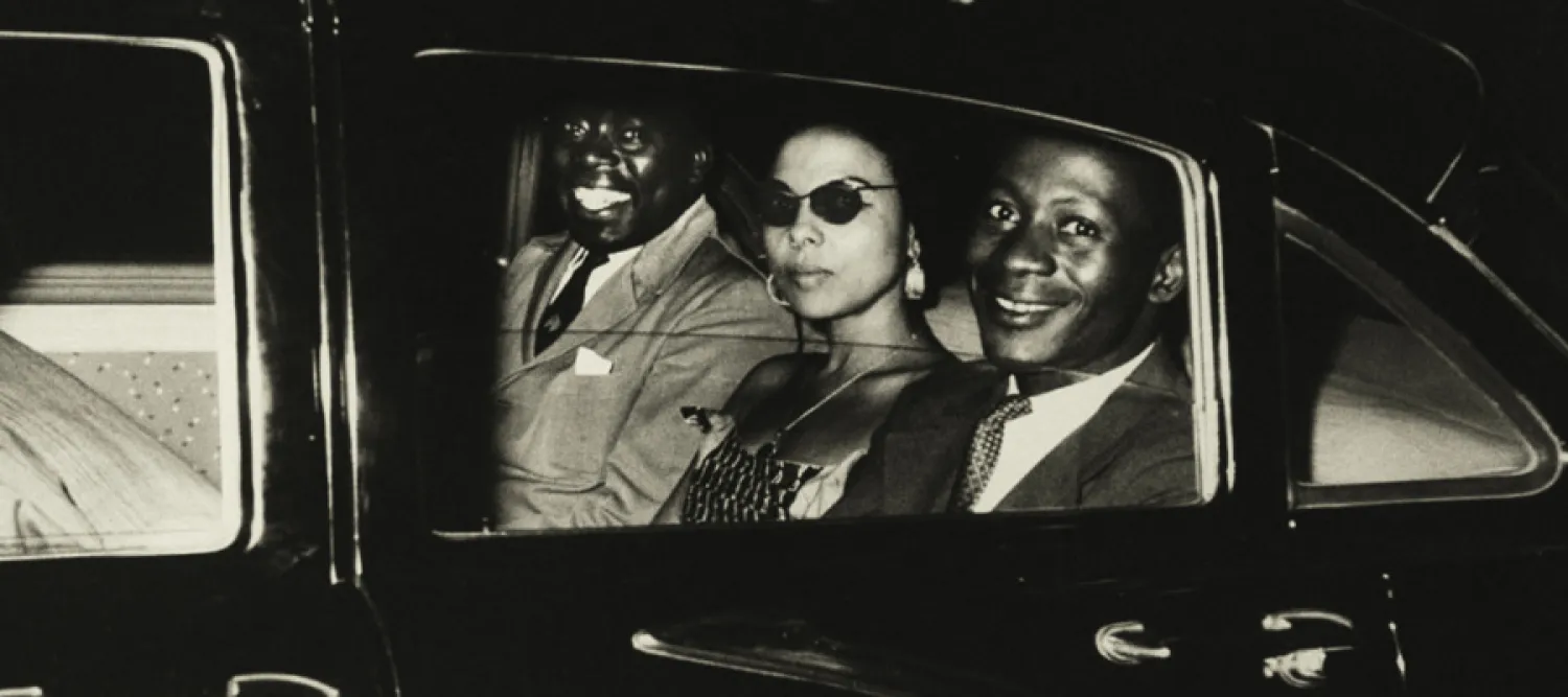 Three people - two men and one woman - sit in the back of a car, looking through a lowered window at the camera.