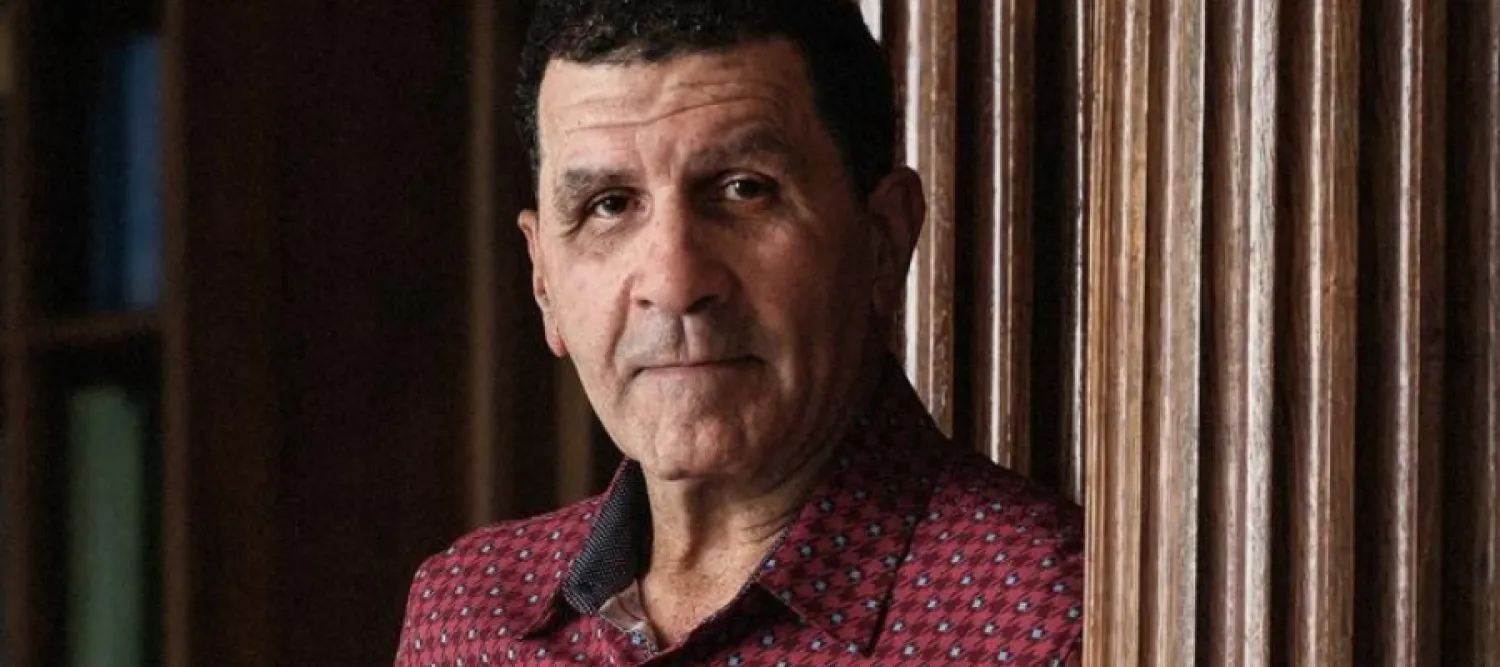 Portrait of a serious-looking man in a red patterned shirt, standing indoors beside dark wooden paneling in warm, subdued light.
