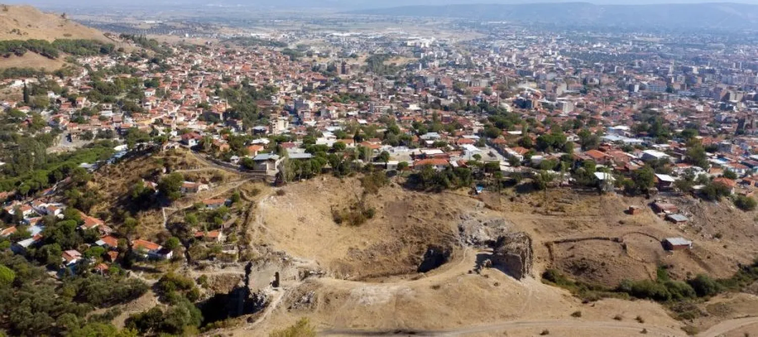 Aerial view of a sprawling hillside town, with dense homes beyond a dry, eroded quarry and winding dirt paths in calm, clear daylight.