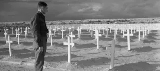 A black and white photograph showing a man looking over a cemetery of white crosses in sand