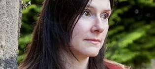 Professor Fiona Edmonds has long dark hair and wears a red coat. She stands against a stone wall, gazing thoughtfully to the side. Lush green foliage forms the background.