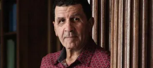 Portrait of a serious-looking man in a red patterned shirt, standing indoors beside dark wooden paneling in warm, subdued light.