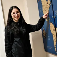 A female student with long dark hair and glasses points to her hometown on the map