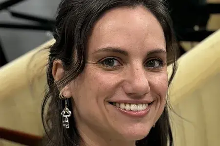 Anna has long dark hair, and looks directly at the camera, smiling