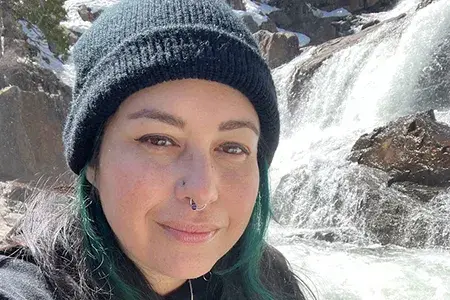 ashley wears a black beanie hat and top, and has a ring through her nose. She is sitting in front of a waterfall and looks directly into the camera.