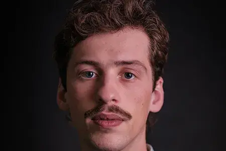 Dominic now, in a professional headshort. He has curly brown hair and a moustache.