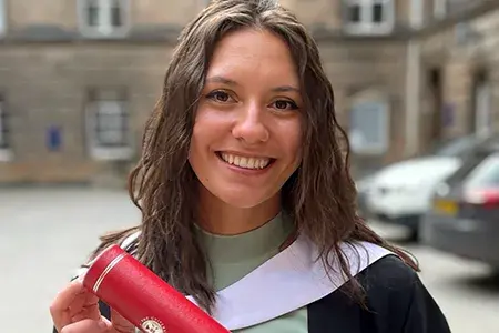 Elise on her graduation day - she wears academic robes and holds a red degree scroll