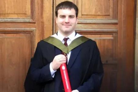 Fergal poses in academic robes, holding his degree scroll