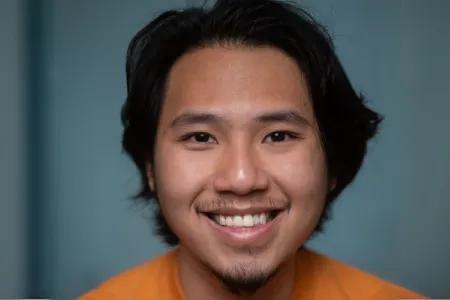 Mai has dark hair and eyes, wears an orange top and smiles broadly at the camera