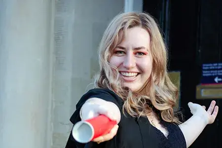 A smiling Tessa on her graduation day points at the camera with a red degree scroll