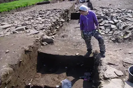 Hannah stands on the edge of an excavation trench, looking down into it