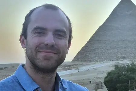 Tristan has dark hair and a beard, and smiles against a backdrop of the pyramids