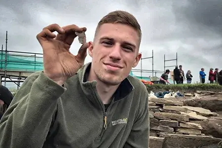 Declan is on an archaeological dig and holds up an artefact