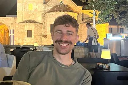 Declan grins broadly at the camera. He has messy hair and a moustache