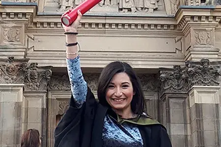 Nicoletta on graduation day. She wears academic robes and holds a loft a graduation scroll.