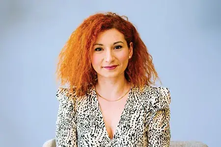 Nicoletta now, in a professional headshot. She has red hair and wears a patterned top.