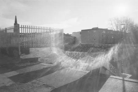 Black-and-white urban skatepark with graffiti-covered concrete ramps, a tall iron fence, and brick apartments; hazy sunlight casts a quiet, moody glow.