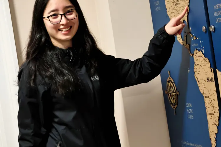 A female student with long dark hair and glasses points to her hometown on the map