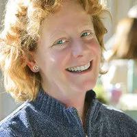 A woman with curly blond hair and a blue sweater smiles at the camera in a brightly lit indoor setting.