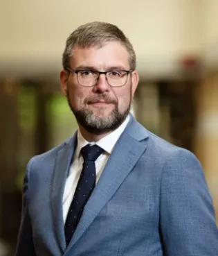 Professor Colin Barr has short fair hair, a beard and wears glasses and a blue suit and navy tie. He stands confidently, and looks directly into the camera.
