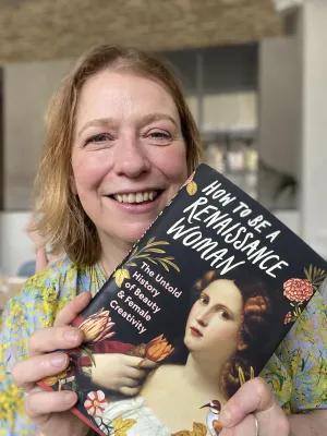 Professor Burke has fair hair, and smiles directly at the camera whilst holding her book titled "How to Be a Renaissance Woman" with a classical painting of a woman on the cover.