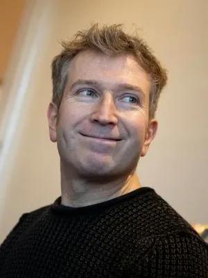 Portrait of a man with short, tousled light-brown hair wearing a black knit sweater, glancing aside with a subtle, relaxed smile against a warm indoor background.