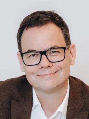 Head-and-shoulders portrait of a smiling man with dark hair and glasses, wearing a brown blazer and white shirt against a light background.