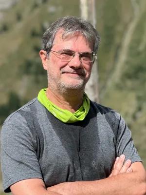 Smiling middle-aged man with glasses and salt-and-pepper hair stands outdoors, arms crossed, wearing a gray shirt and bright green neck gaiter.