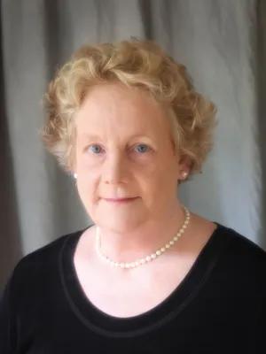 Studio portrait of a mature woman with short, softly curled blonde hair, wearing a pearl necklace and black top against a gray draped backdrop; her expression is calm and warm.