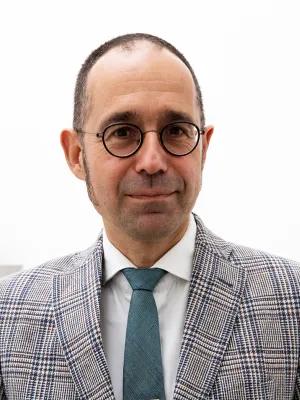 A bespectacled man in a checked blazer, white shirt, and teal tie faces the camera against a bright white background, wearing a calm, approachable expression.