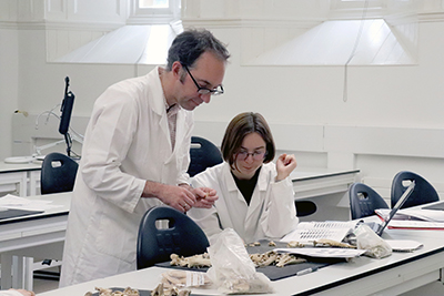 HCA A tutor and student wearing white lab coats study finds in the School archaeology teaching lab made up of white benches and chairs.